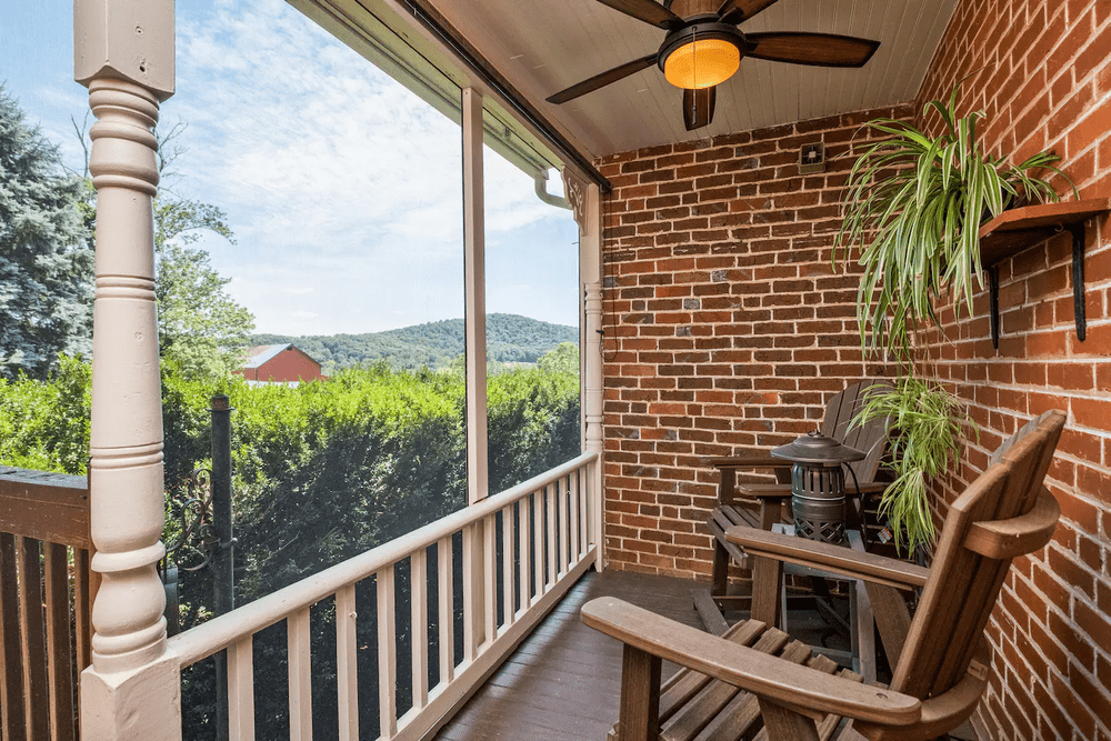 Cozy screened porch featuring wooden rocking chairs, a decorative lantern, and a vibrant green plant, with a scenic view of rolling hills and blue skies in the background.