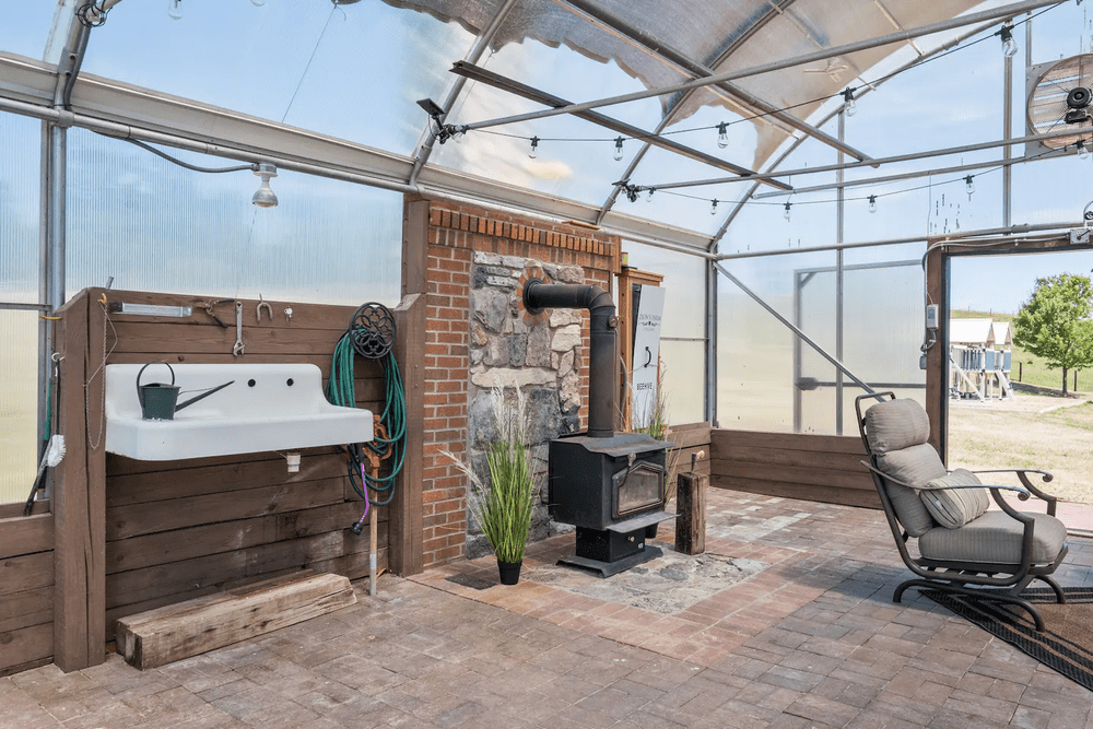 Interior view of a greenhouse featuring a vintage sink, a wood-burning stove, and a comfortable rocking chair, with a brick wall and large windows allowing natural light. The space is designed for gardening and relaxation, showcasing a blend of rustic and functional elements.