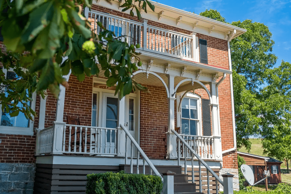 Historic brick house with a welcoming porch, featuring white railings and steps, surrounded by lush greenery and a clear blue sky.