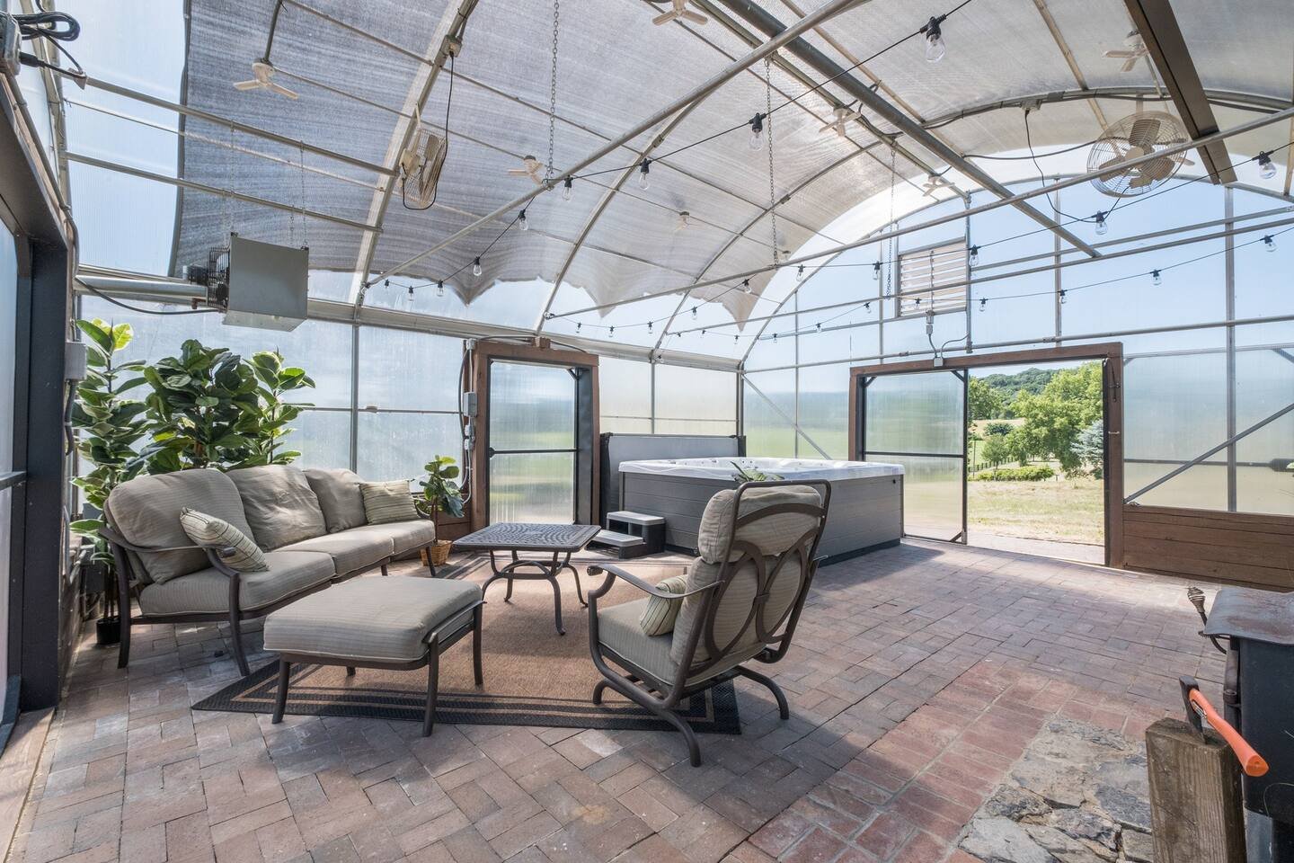 Spacious indoor greenhouse featuring a cozy seating area with a sofa and armchair, a hot tub, and lush greenery. Natural light floods the space through transparent walls and ceiling, creating a serene atmosphere. Ideal for relaxation and enjoying nature indoors.